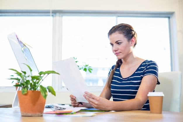 A young woman sitting at a desk, looking at a laptop with a spreadsheet open. She has a thoughtful expression, and there's a piggy bank and some books on finance on the desk around her, suggesting she is learning about personal finance.